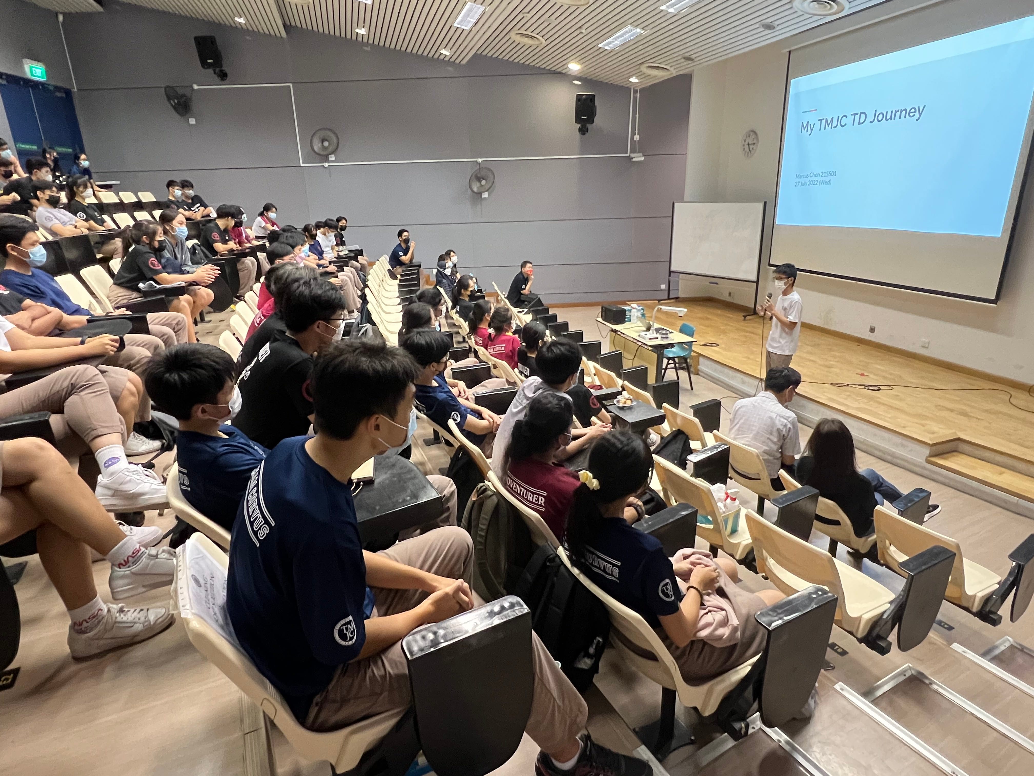 Students sit in a lecture hall watching a presenter standing near a screen showing "My TMJC TD Journey."
